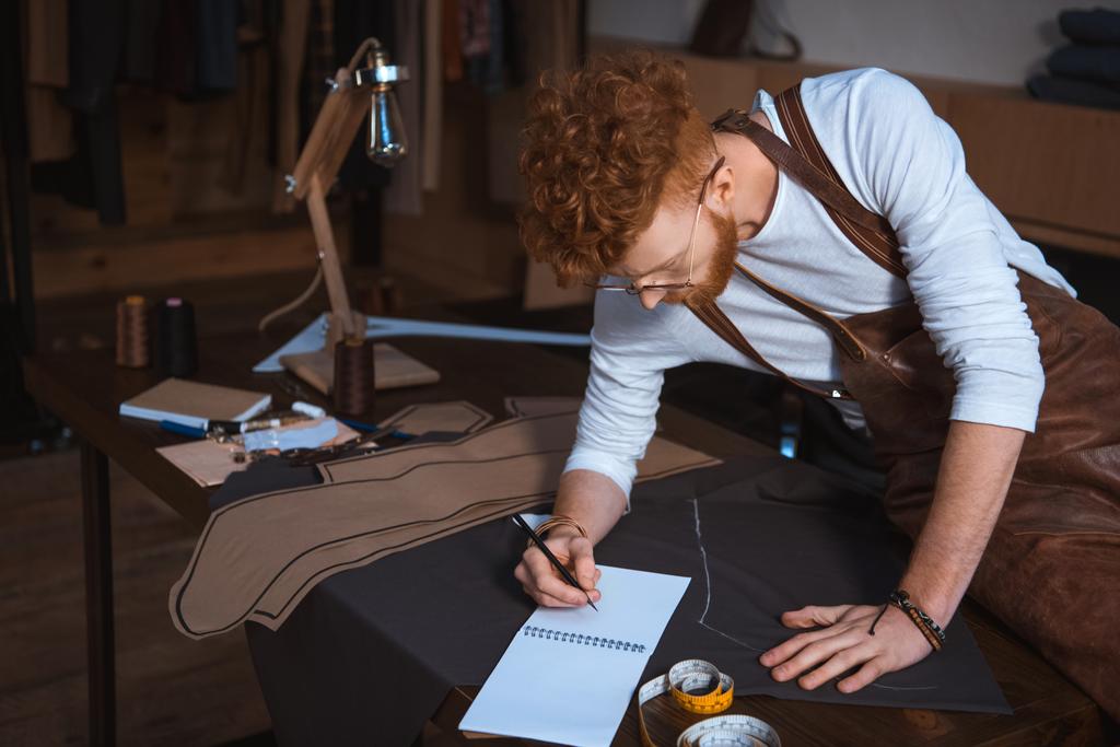 młody mężczyzna projektant mody w fartuch i okulary, robić notatki w notesie  - Zdjęcie, obraz