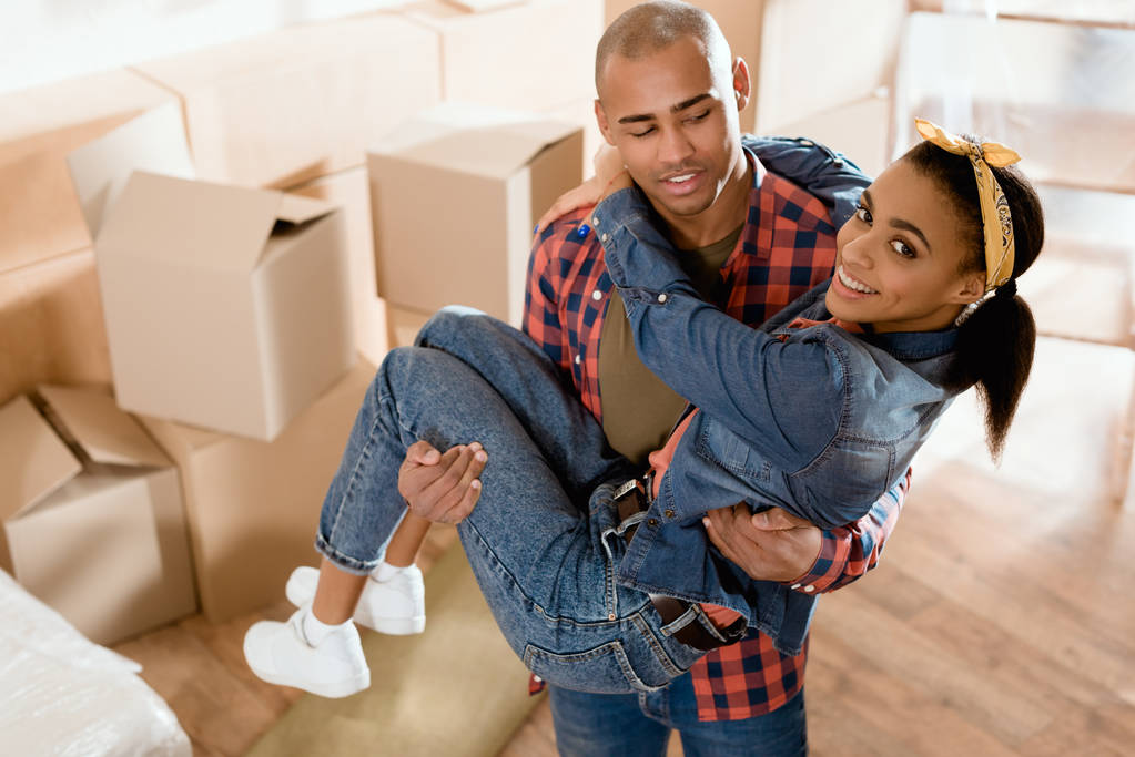 african american boyfriend holding on hands his girlfriend in new apartment with cardboard boxes - Photo, Image
