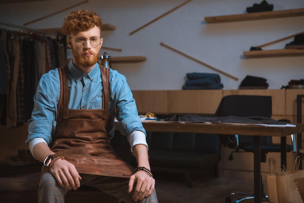 seriöser männlicher Modedesigner in Schürze und Brille sitzt in der Werkstatt - Foto, Bild