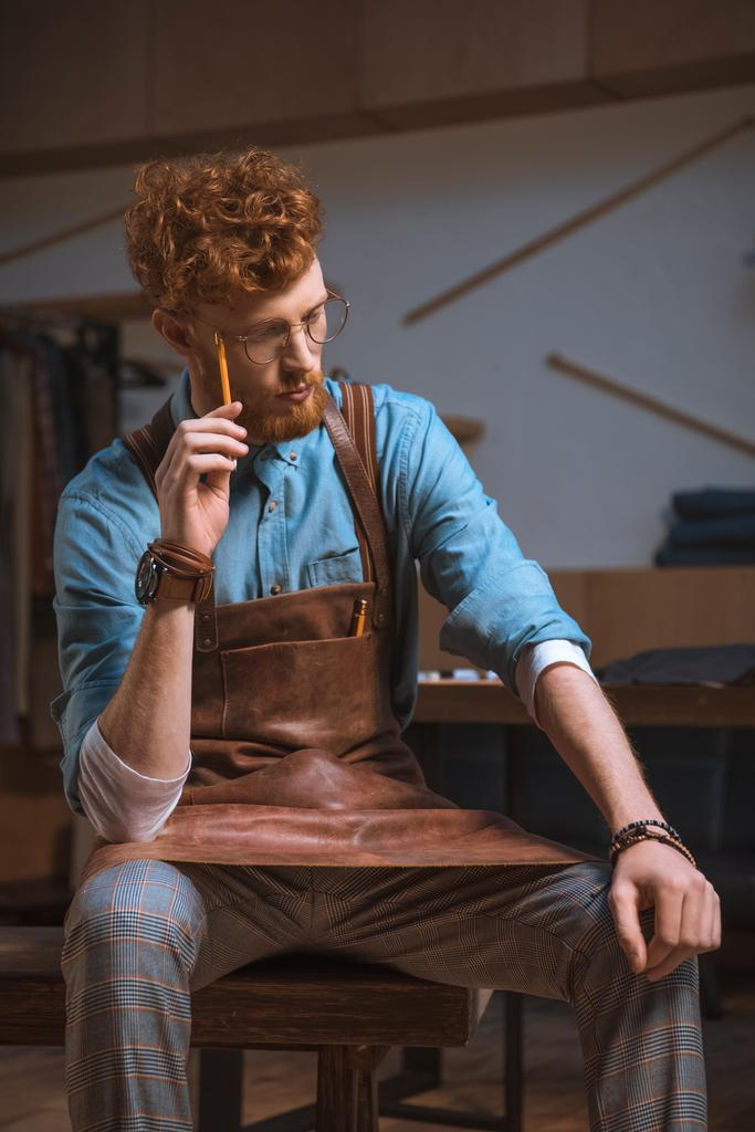 συλλογισμένος νεαρά αρσενικά σχεδιαστής μόδας στην ποδιά και γυαλιά, κάθεται και σκέφτεται με μολύβι στο χέρι - Φωτογραφία, εικόνα