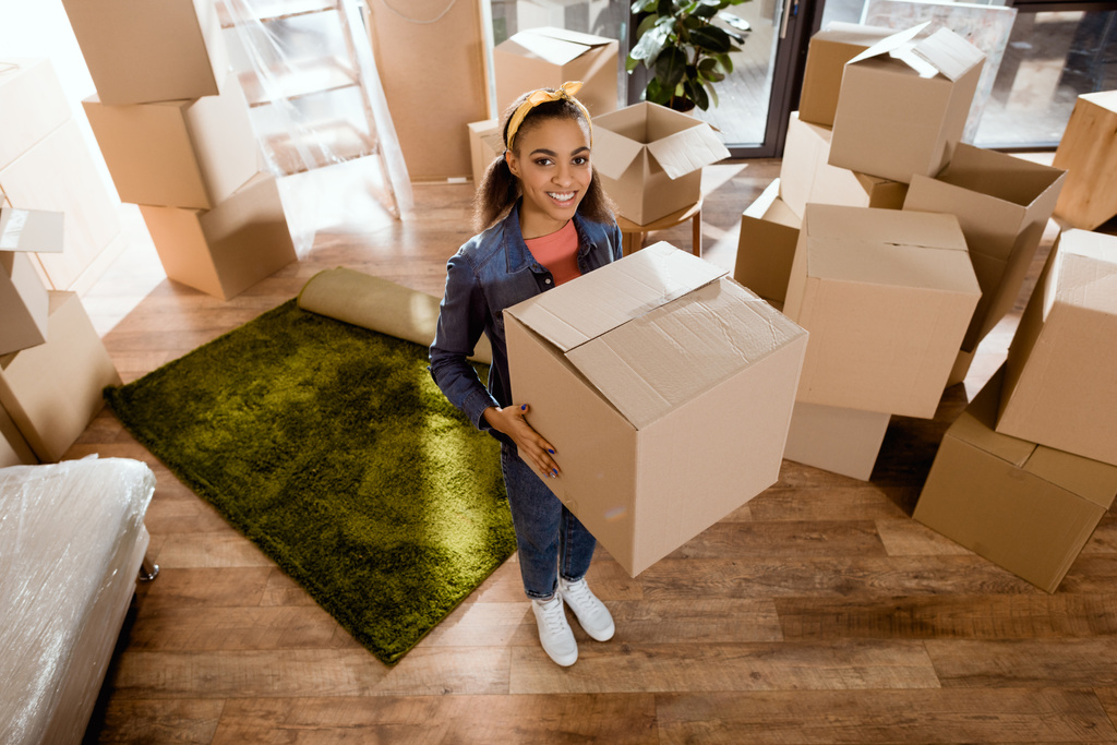 молодая улыбающаяся африканская американка держит картонную коробку и переезжает в новый дом
 - Фото, изображение