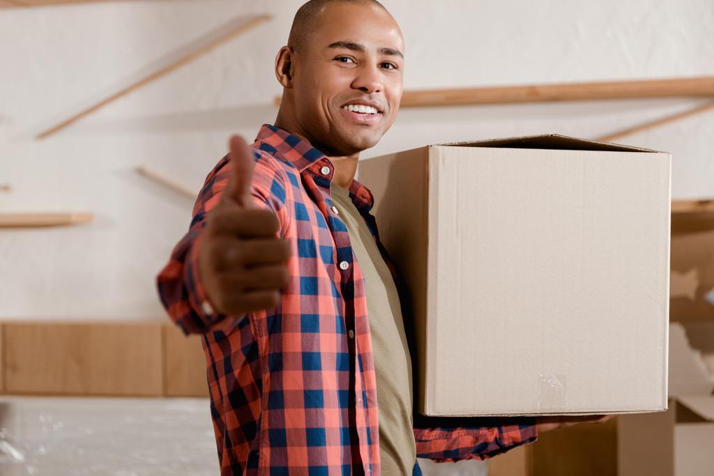 junger afrikanisch-amerikanischer Mann mit erhobenem Daumen hält Pappschachtel im neuen Zuhause - Foto, Bild