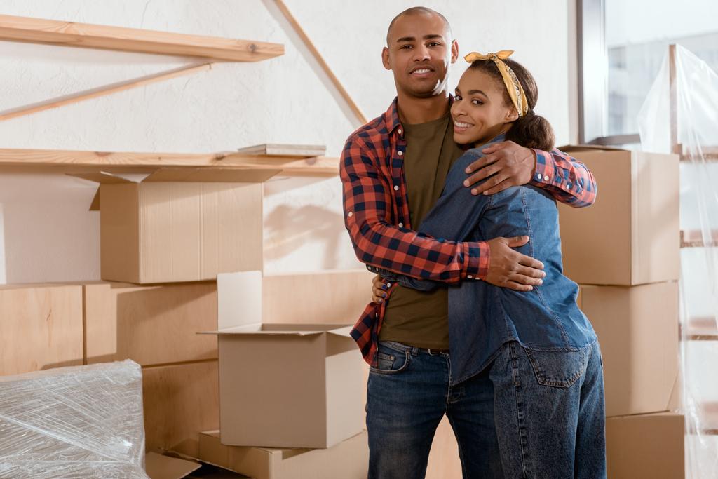 Gelukkig Afrikaanse Amerikaanse echtpaar knuffelen in nieuw appartement met kartonnen dozen - Foto, afbeelding