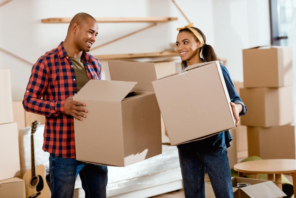 lachende Afrikaanse Amerikaanse echtpaar houden van kartonnen dozen en bewegen naar nieuwe thuis - Foto, afbeelding