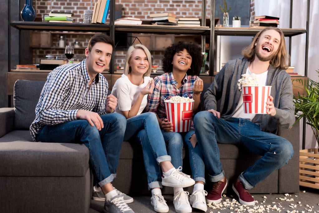 amigos multiculturales riendo viendo la televisión en el sofá con palomitas de maíz
 - Foto, Imagen