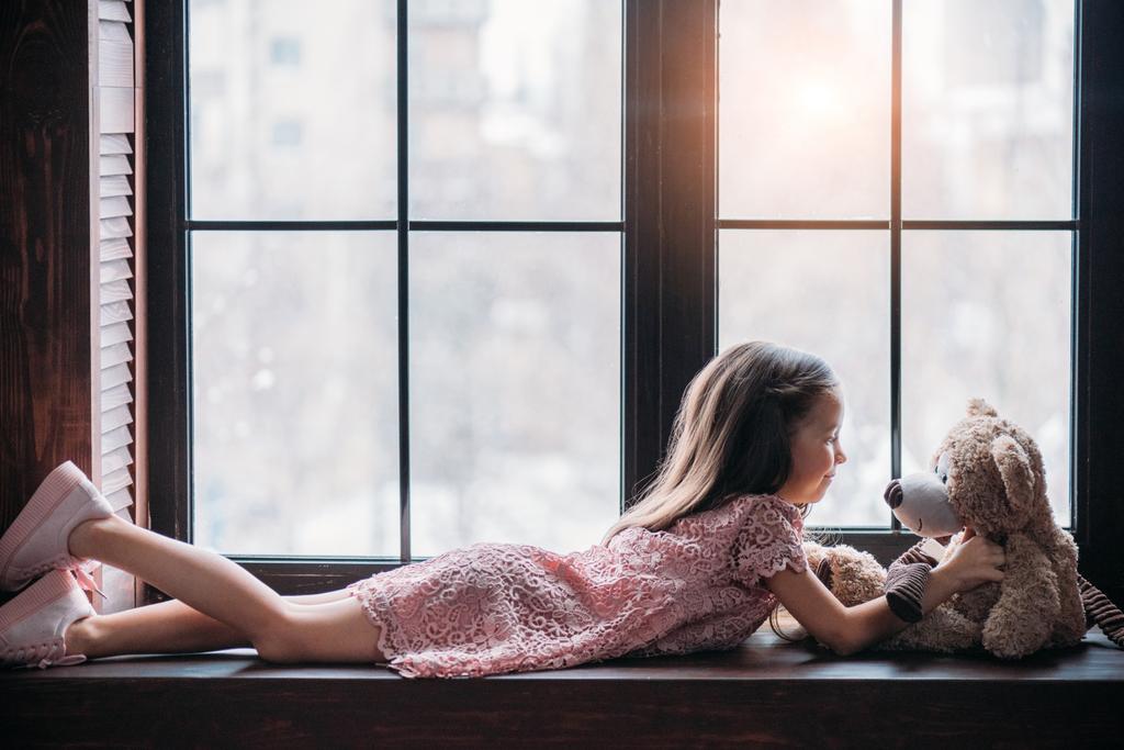side view of little child playing with teddy bear while lying on windowsill - Photo, Image