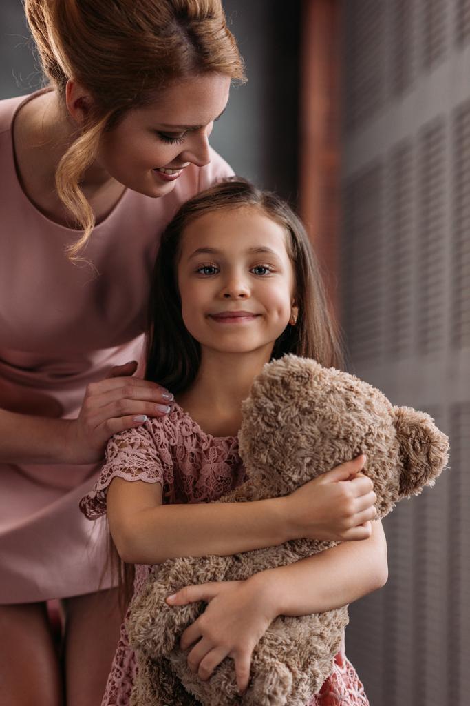 junge schöne Mutter umarmt ihre Tochter, während sie einen Teddybär hält - Foto, Bild