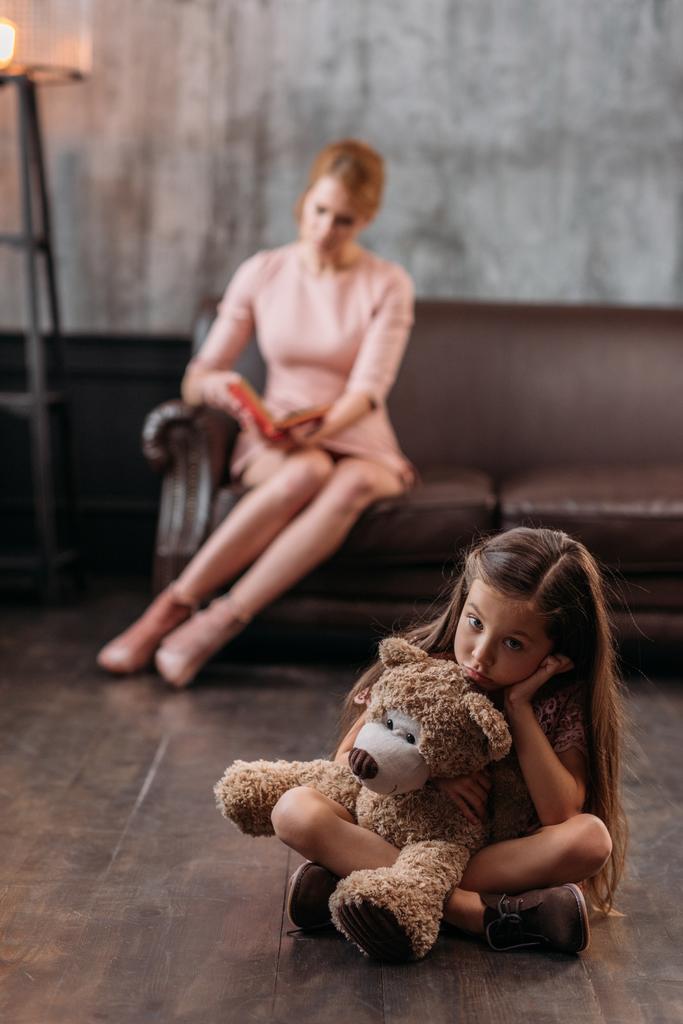 weinig depressief kind zittend op de vloer met teddy beer terwijl haar moeder zittend op de Bank- en leesboek - Foto, afbeelding