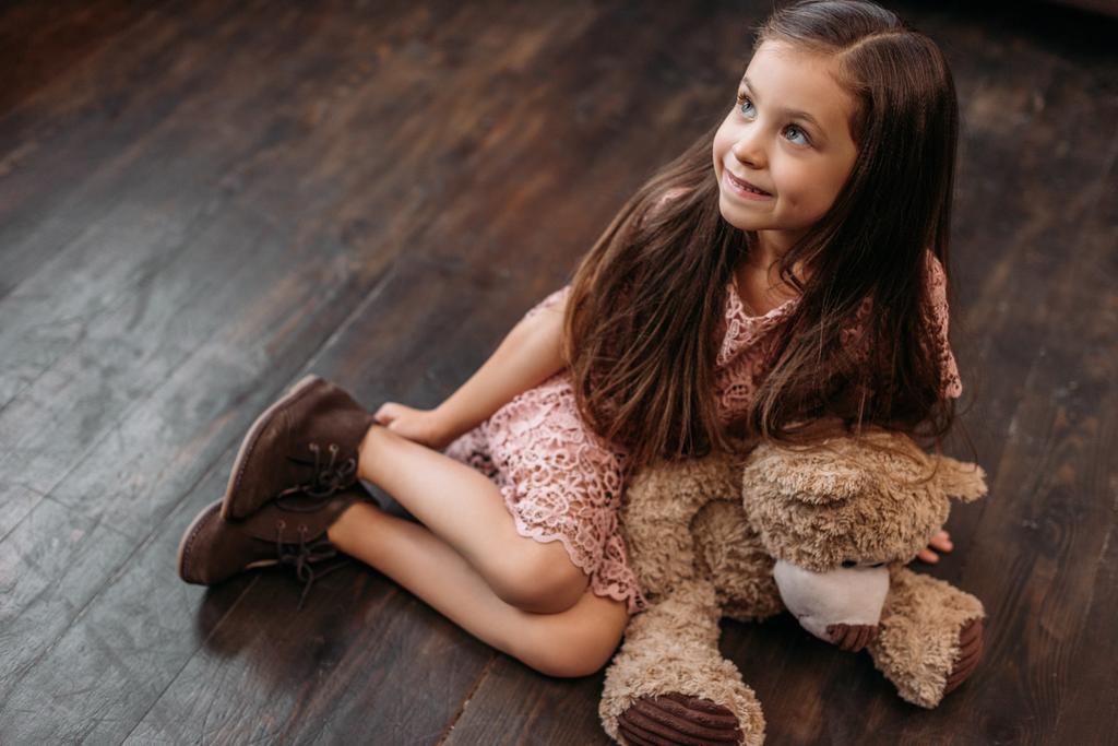 suloinen pieni lapsi istuu lattialla nallekarhun kanssa
 - Valokuva, kuva