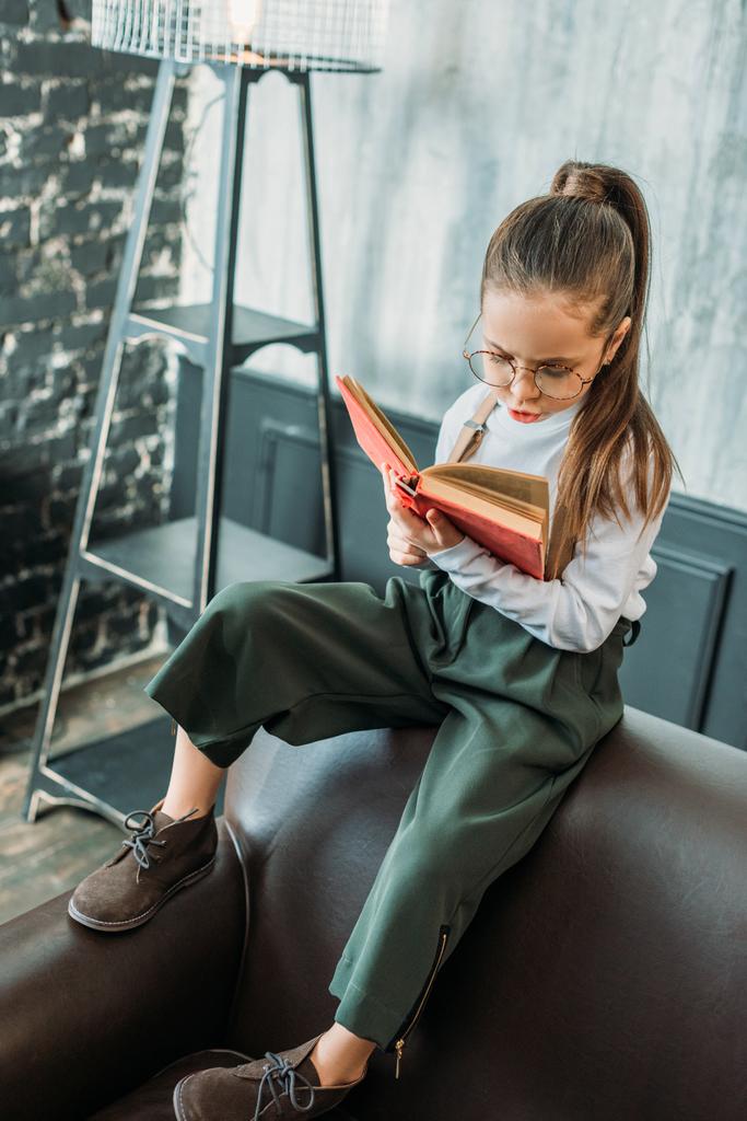 Концентрированный маленький ребенок читает книгу, сидя на диване
 - Фото, изображение