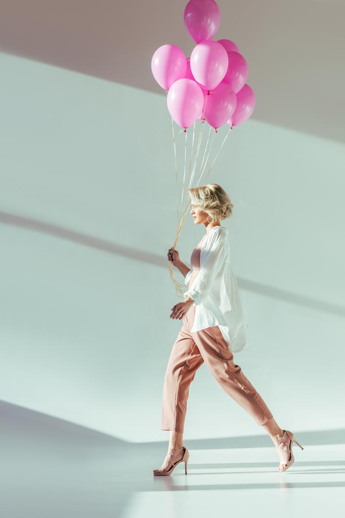 Zijaanzicht van mooie stijlvolle jongedame lopen met roze ballonnen op grijs - Foto, afbeelding