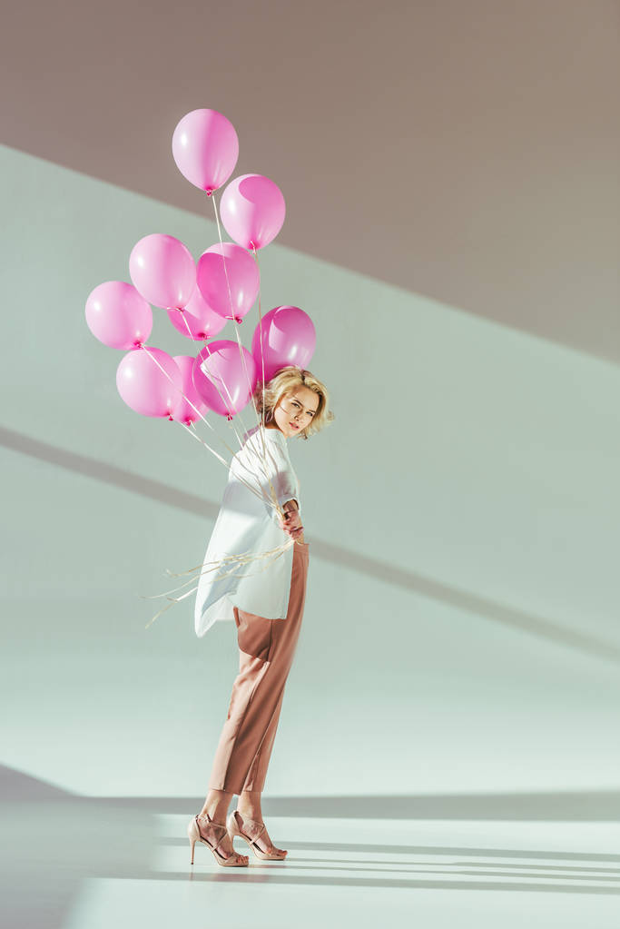 gyönyörű divatos fiatal nő gazdaság rózsaszín léggömbök és látszó-on fényképezőgép-a szürke - Fotó, kép