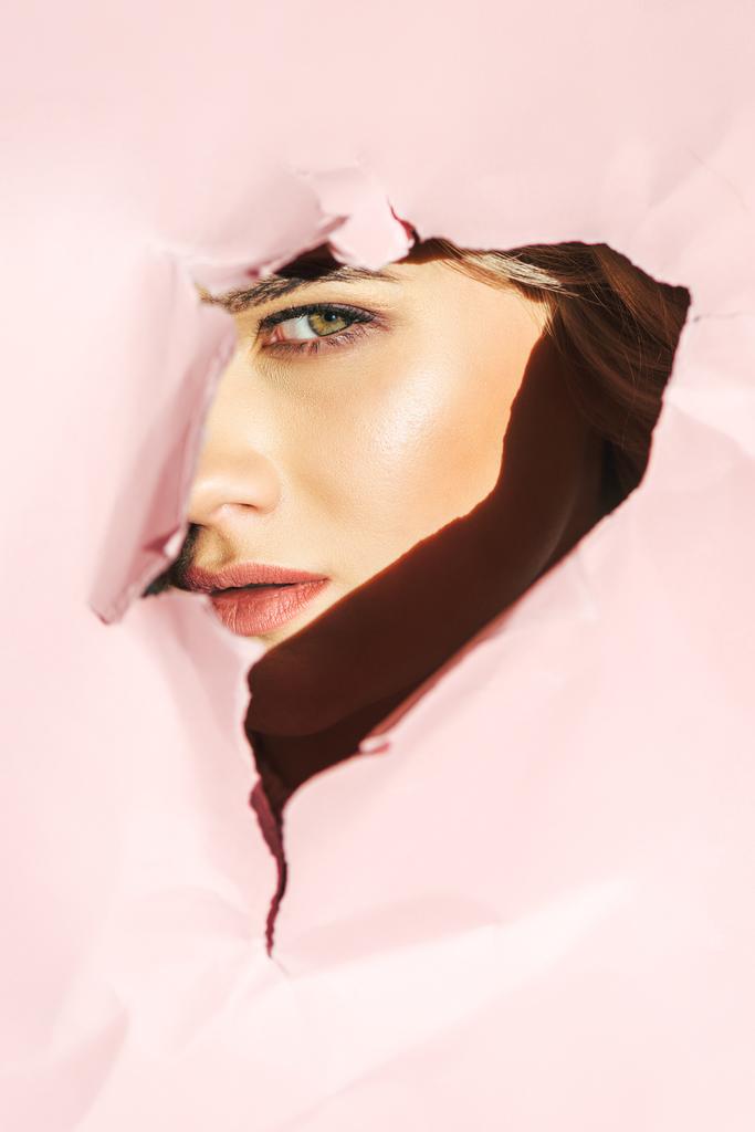 attraktive junge Frau blickt durch zerrissenes rosa Papier in die Kamera   - Foto, Bild