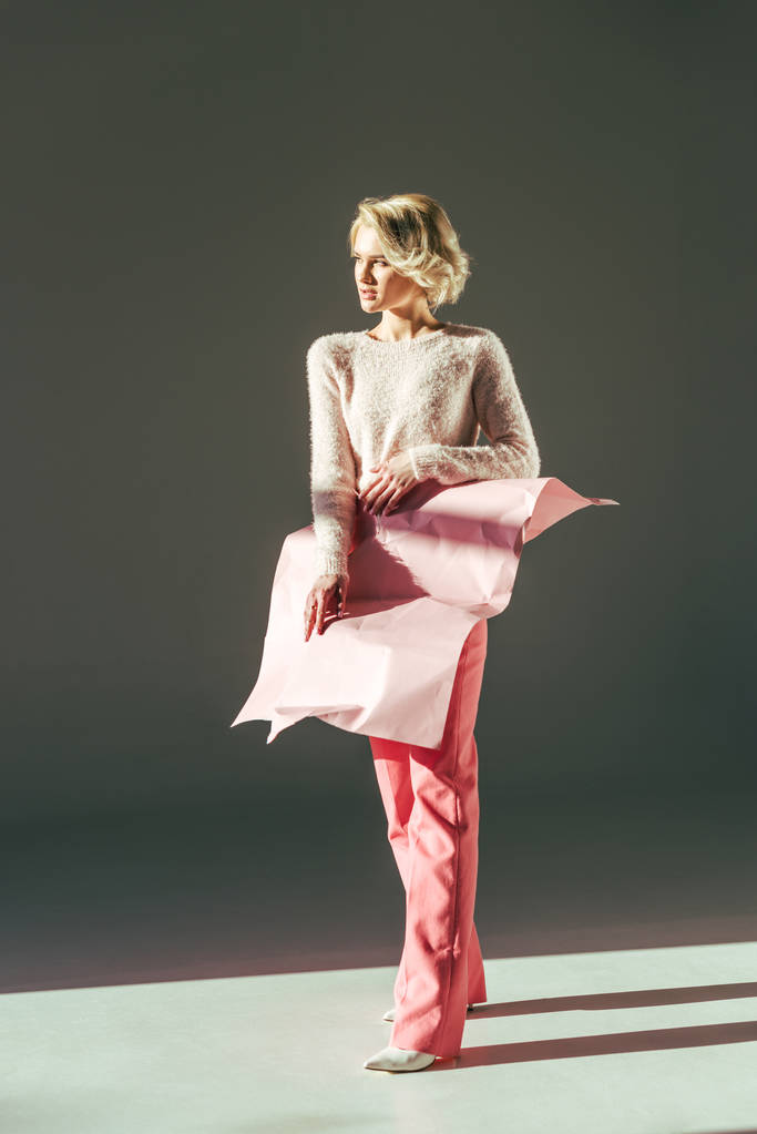 modieuze jonge blonde vrouw met roze verfrommeld papier aan taille kijken weg in studio - Foto, afbeelding