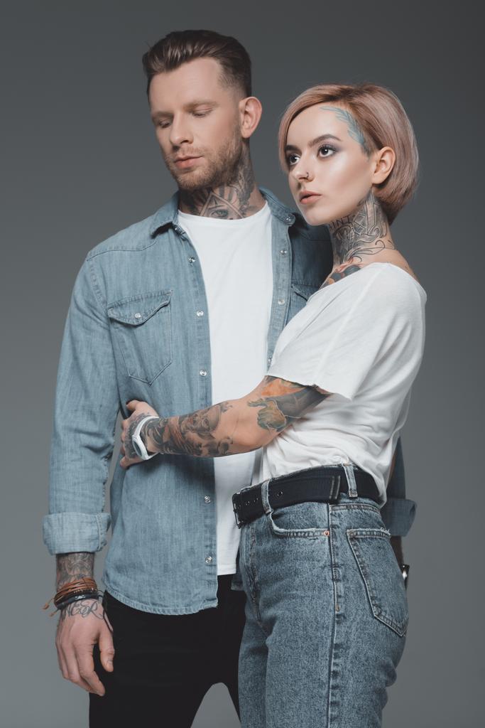 schönes stylisches junges Paar mit Tätowierungen, die sich umarmen und isoliert auf grau wegschauen - Foto, Bild