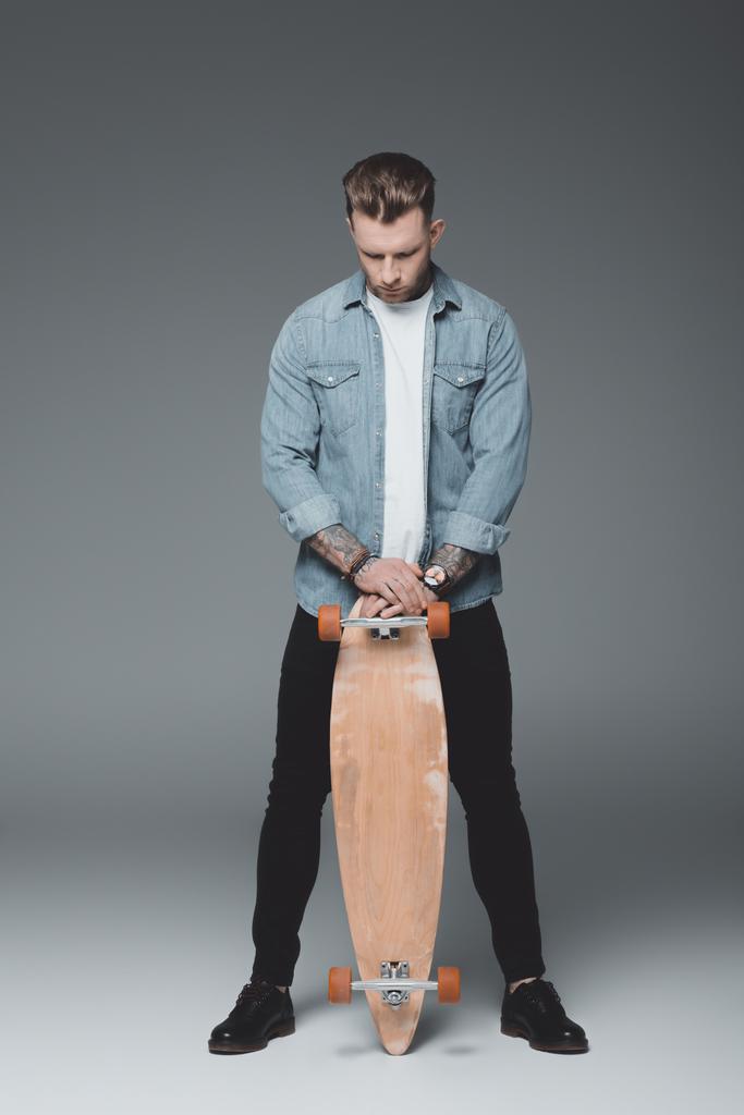 Ganzkörperansicht eines hübschen jungen tätowierten Mannes, der mit Skateboard auf grau steht - Foto, Bild
