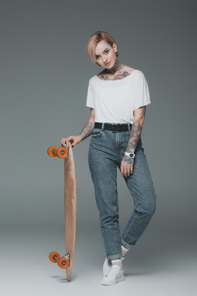 mooie getatoeëerd meisje permanent met skateboard en camera kijken op grijs  - Foto, afbeelding