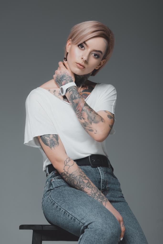beautiful tattooed girl in white t-shirt sitting on stool and looking at camera isolated on grey - Photo, Image