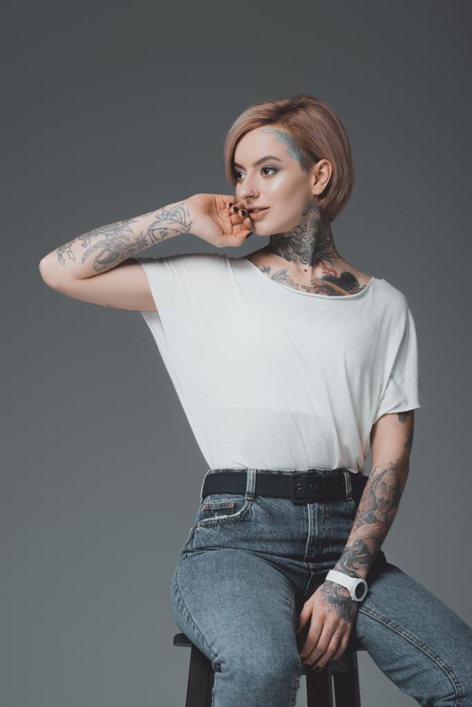 beautiful smiling young woman with tattoos sitting on stool and looking away isolated on grey - Photo, Image