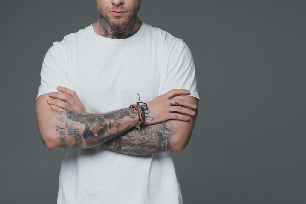 bijgesneden schot van jonge man met tatoeages permanent met gekruiste armen geïsoleerd op grijs - Foto, afbeelding