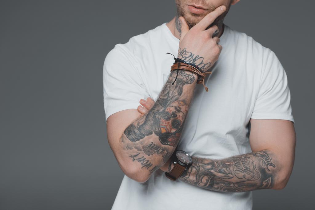 обрезанный снимок стильного молодого татуированного мужчины в белой футболке, стоящего с рукой на подбородке, изолированного на сером
 - Фото, изображение