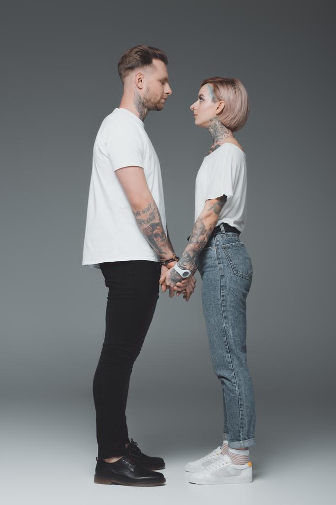 schöne stylische junge tätowierte Paar in weißen T-Shirts Händchen haltend und einander anschauend auf grau - Foto, Bild