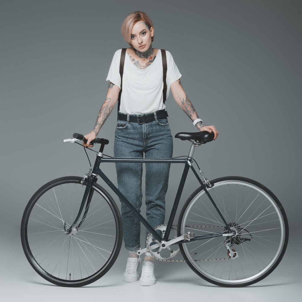 piękna stylowa dziewczyna z tatuażami, stojąc z roweru i patrząc na kamery na szarym tle  - Zdjęcie, obraz