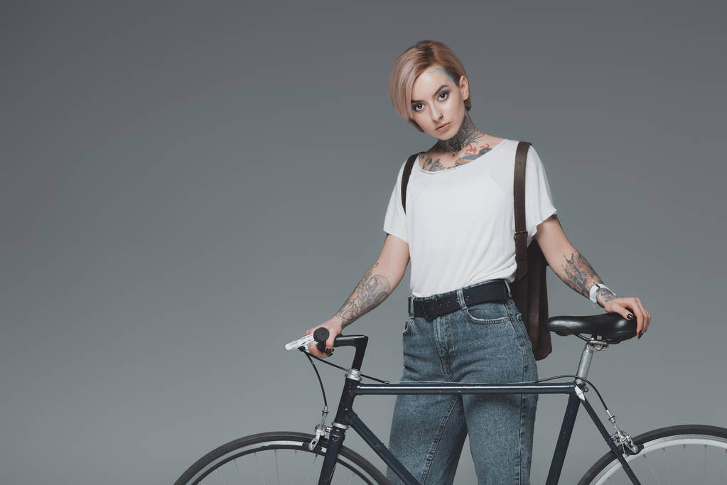 schönes tätowiertes Mädchen, das mit Fahrrad steht und isoliert auf grau in die Kamera schaut  - Foto, Bild