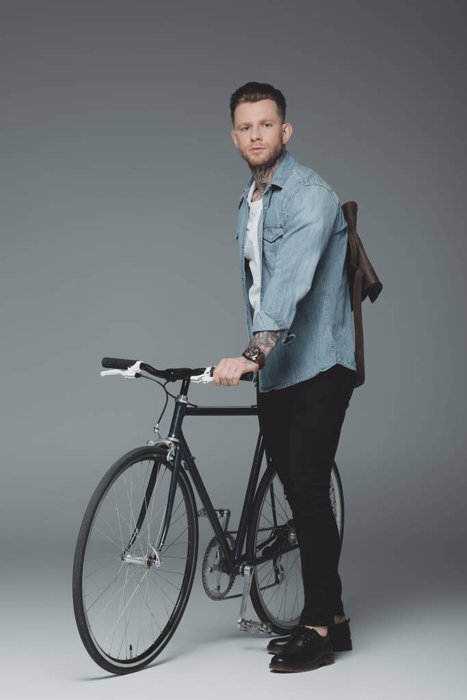 повнометражний вид на стильного молодого чоловіка з татуюваннями, що стоять з велосипедом і дивляться на камеру на сірому
 - Фото, зображення