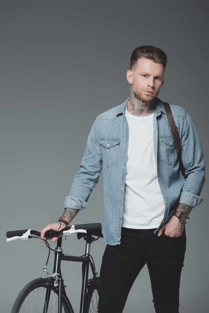 красивый молодой человек с плечевой сумкой и велосипедом, стоящий и смотрящий на камеру, изолированную на сером
 - Фото, изображение