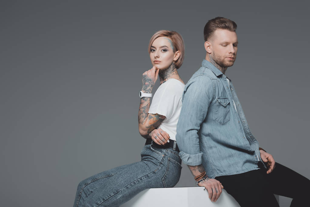 elegante joven pareja tatuada sentado espalda con espalda y mirando a la cámara aislada en gris
  - Foto, imagen