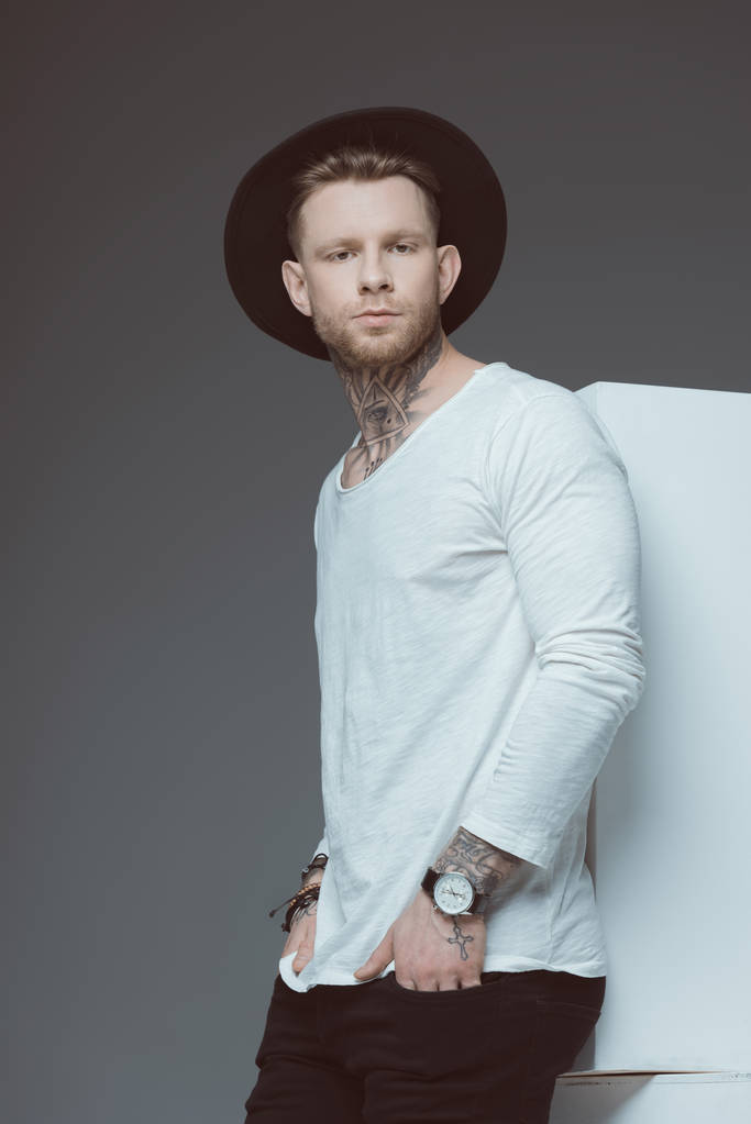 joven hombre tatuado con estilo en sombrero negro de pie con las manos en los bolsillos y mirando a la cámara aislada en gris
 - Foto, Imagen