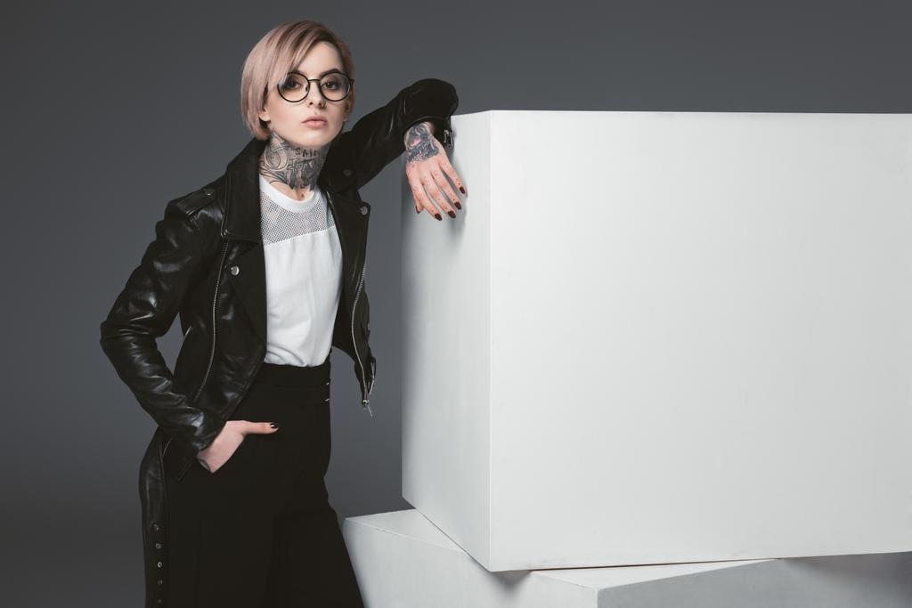 beautiful young tattooed woman in leather jacket leaning at white cubes and looking at camera isolated on grey - Photo, Image