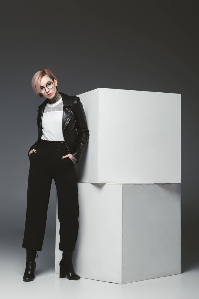 fashionable young woman in leather jacket and eyeglasses standing with hands in pockets and looking at camera in studio - Photo, Image