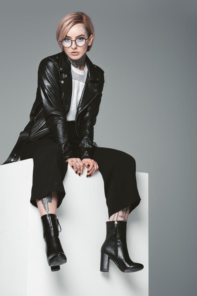 stylische junge Frau mit Tätowierungen in Lederjacke und Blick in die Kamera isoliert auf grau - Foto, Bild