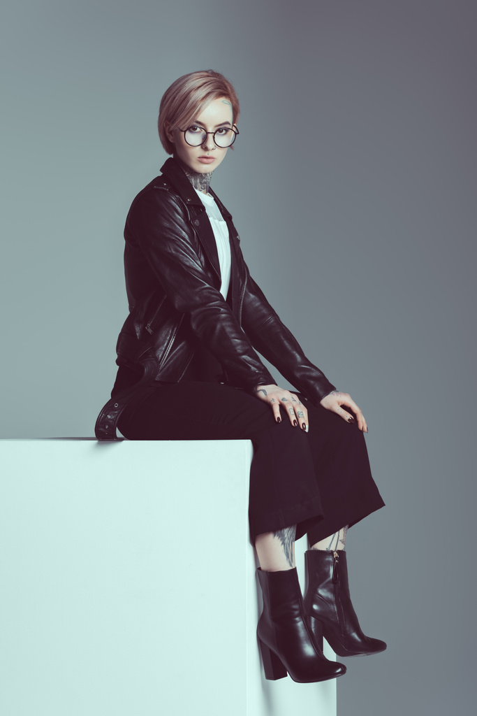 beautiful tattooed girl in leather jacket and spectacles sitting and looking at camera isolated on grey - Photo, Image