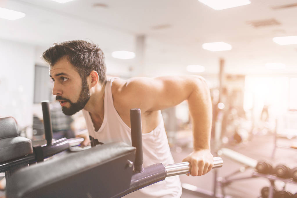 Muscular bearded man during workout in the gym. A portrait of a focused athletic male model in white clothes doing dips on parallel bars . Indoor fitness - Photo, Image