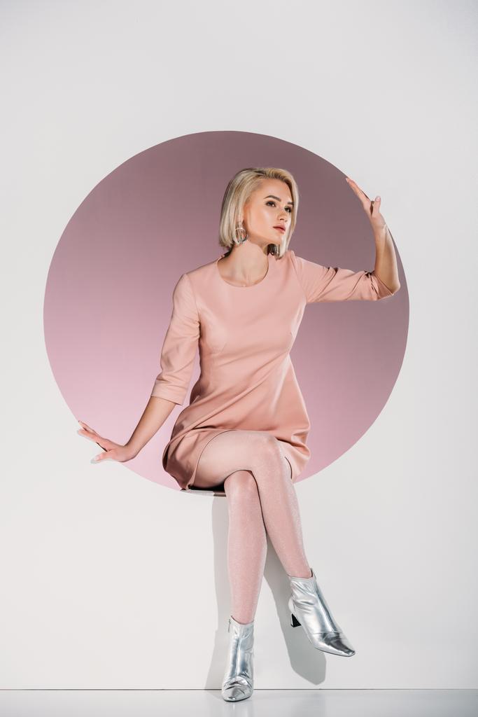 beautiful stylish blonde woman in dress and shiny shoes sitting in hole and looking away on grey  - Photo, Image