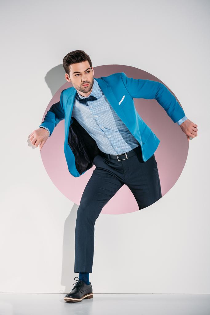 guapo joven con estilo en traje y corbata de lazo mirando hacia otro lado mientras camina a través del agujero en gris
 - Foto, imagen