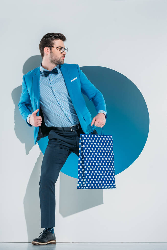 handsome stylish young man with shopping bag looking away while stepping through hole on grey - Photo, Image