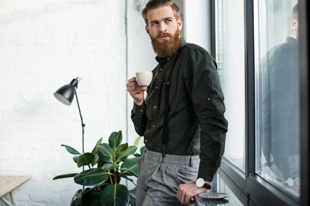 бородатый бизнесмен стоит с чашкой кофе и смотрит в сторону
 - Фото, изображение