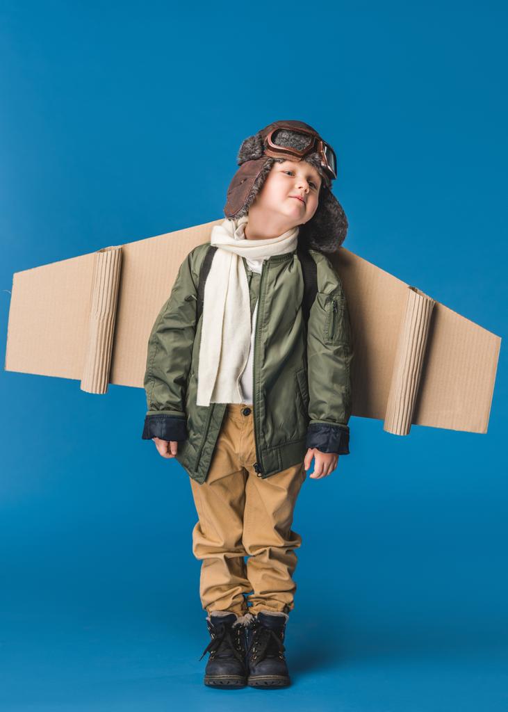 χαριτωμένο μικρό αγόρι στο πιλοτικό κοστούμι με χαρτί αεροπλάνο πτέρυγα απομονώνονται σε μπλε - Φωτογραφία, εικόνα