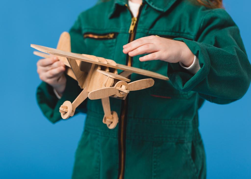 bijgesneden schot van kind in piloot kostuum met houten vliegtuig speelgoed geïsoleerd op blauw - Foto, afbeelding