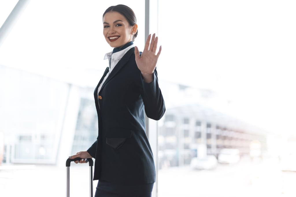 привлекательная молодая стюардесса с чемоданом, машущим перед камерой в аэропорту
 - Фото, изображение