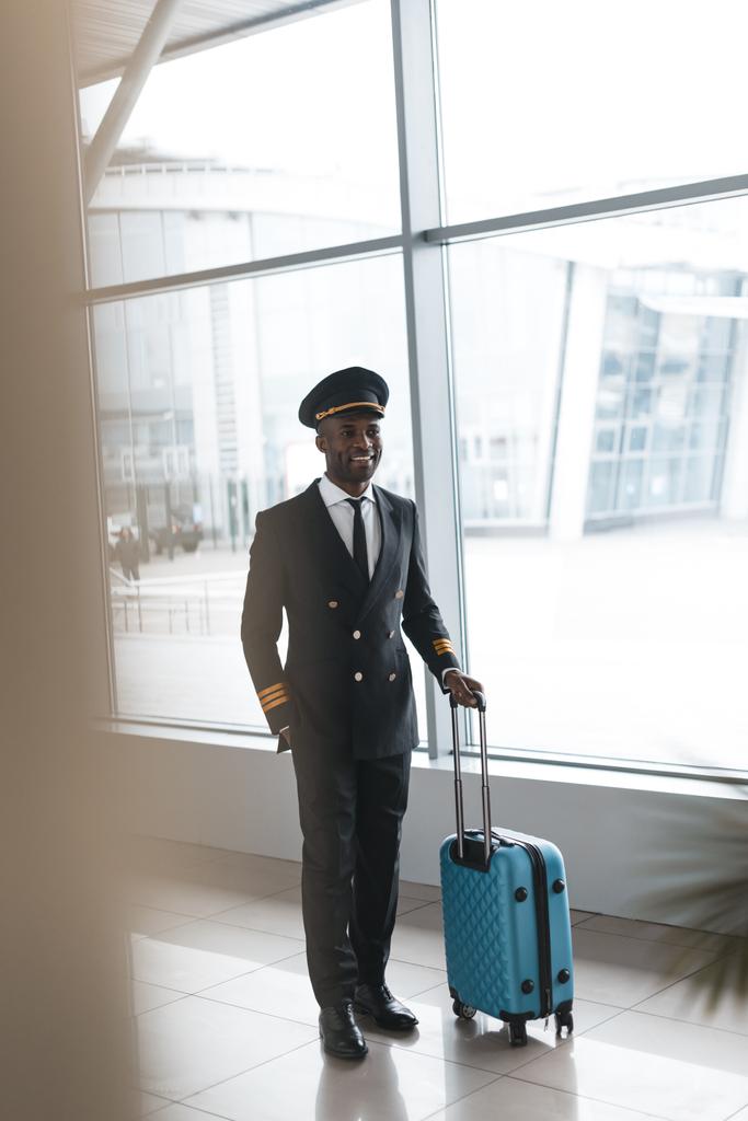 mosolygó fiatal pilóta profi egységes bőrönddel repülőtéren - Fotó, kép