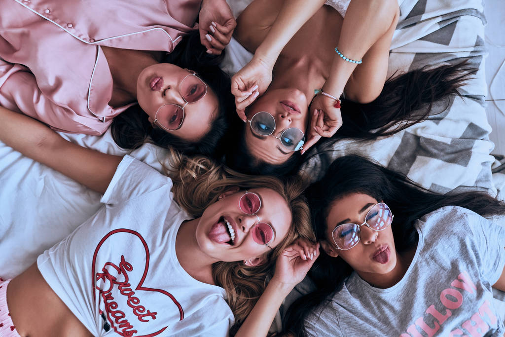 Вид сверху на четырех веселых молодых женщин в экипировке, улыбающихся и делающих лицо, лежа на кровати
 - Фото, изображение