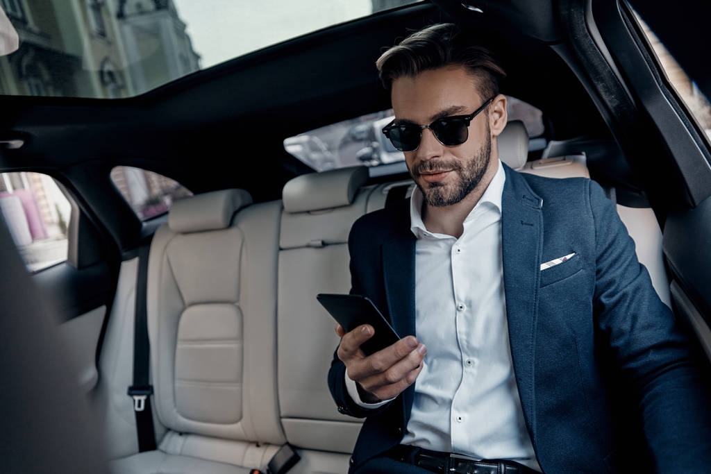Knappe jonge man in pak zijn slimme telefoon kijken zittend in de auto - Foto, afbeelding