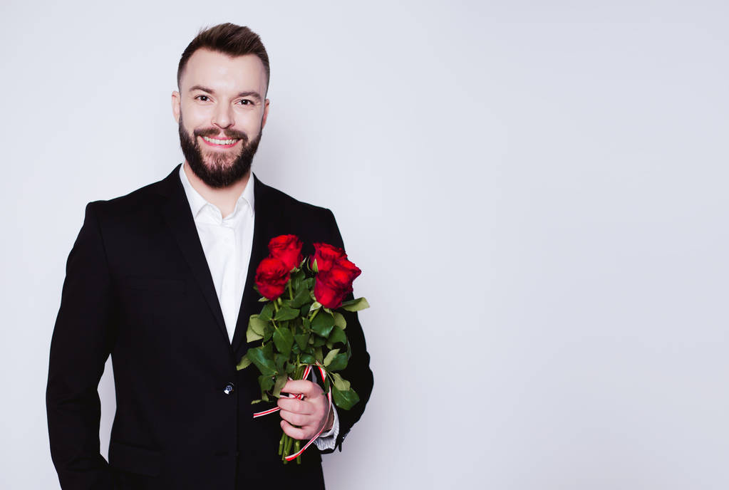 bel homme souriant dans un élégant costume noir avec des roses rouges dans les mains regarde la caméra sur le fond blanc isolé
. - Photo, image