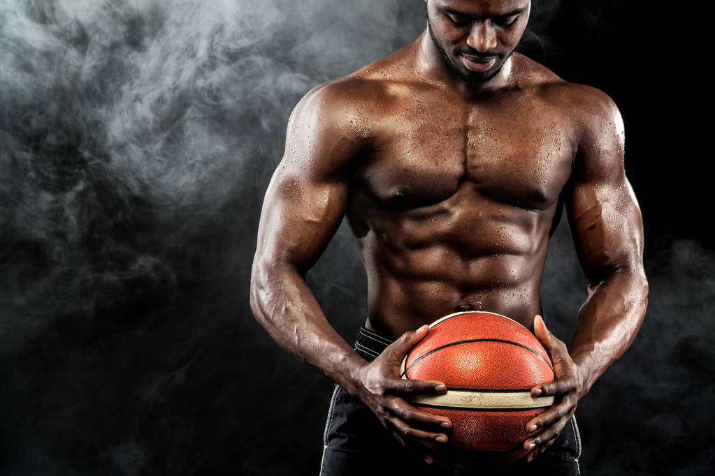 Πορτρέτο της αφροαμερικάνικης αθλητή, μπασκετμπολίστας με μια μπάλα πάνω από το μαύρο φόντο. FIT νεαρός άνδρας σε αθλητικά ρούχα κρατώντας την μπάλα. - Φωτογραφία, εικόνα