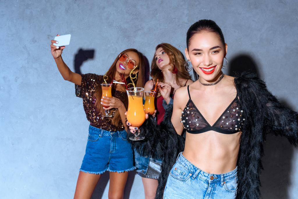 s úsměvem elegantní Asijské žena hospodářství skla s koktejl, zatímco přátelé pití koktejlů a pořizování selfie s smartphone za v strana  - Fotografie, Obrázek
